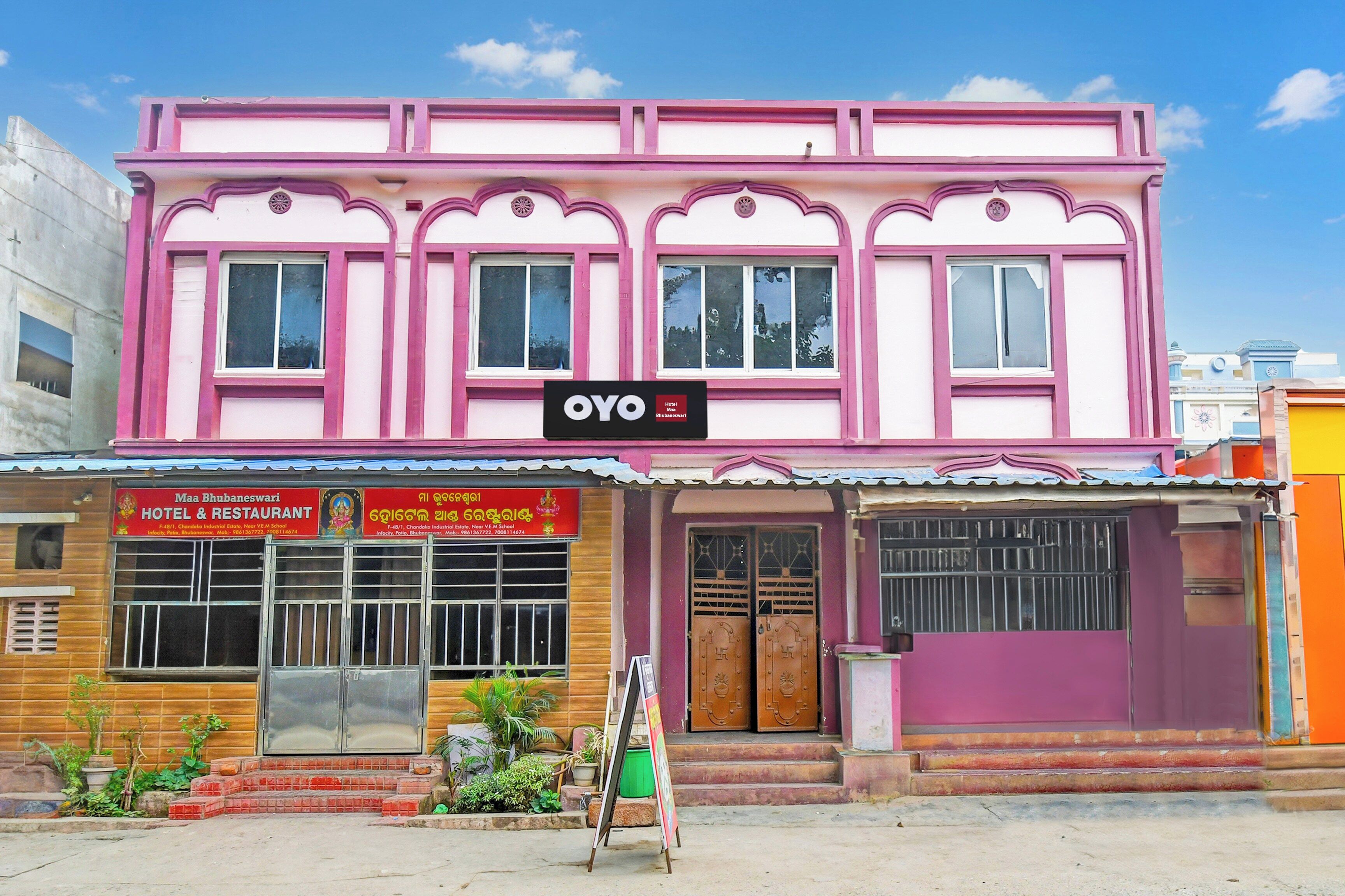 A pink building with a sign for 'OYO Hotel' and another sign for 'Maa Bhubaneswari Hotel & Restaurant' with contact information.