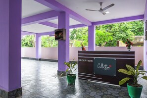 Un área de recepción con un mostrador identificado como 'Collection Signature Stays by G&J'. Hay plantas en macetas a ambos lados del mostrador y un ventilador de techo arriba.