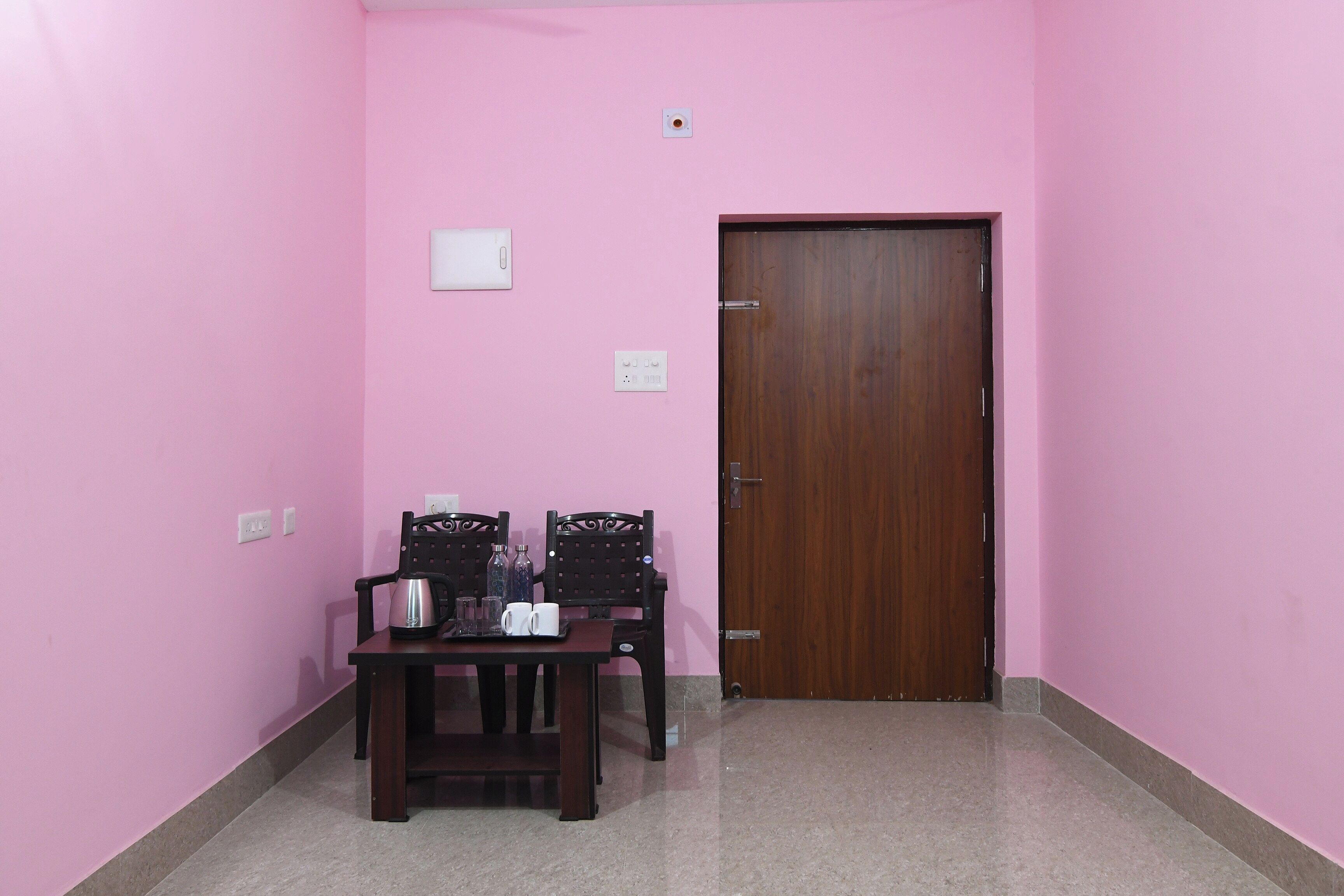 A room with pink walls, a wooden door, a table with chairs, and a small table with a kettle and cups.