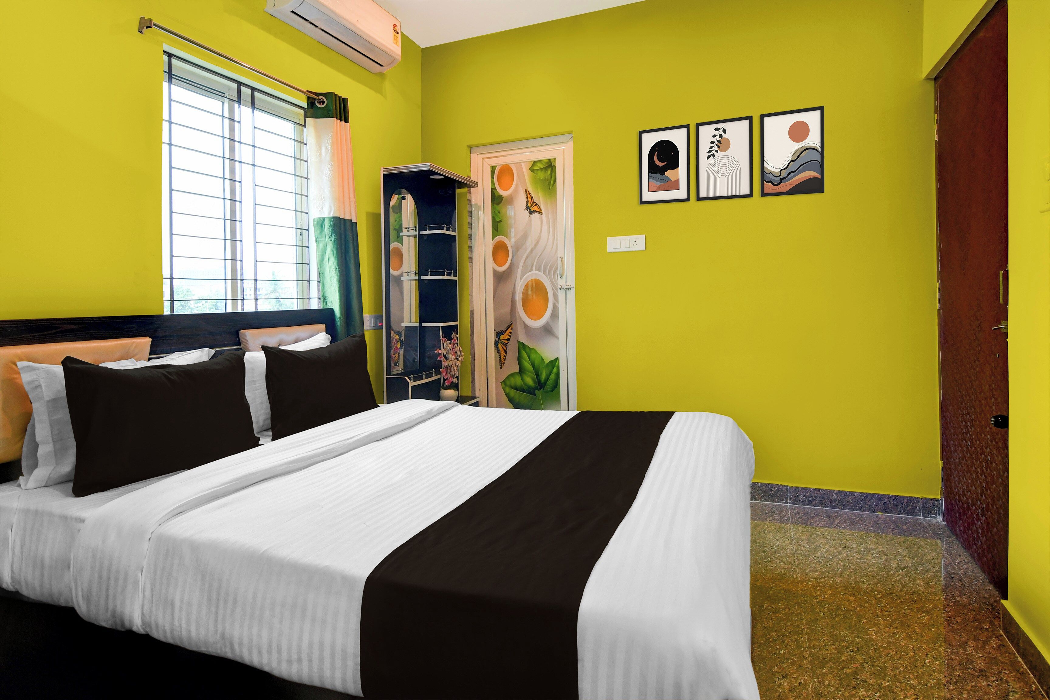 A hotel room with a bed, a window with blinds, a door with a mirror, and wall art.