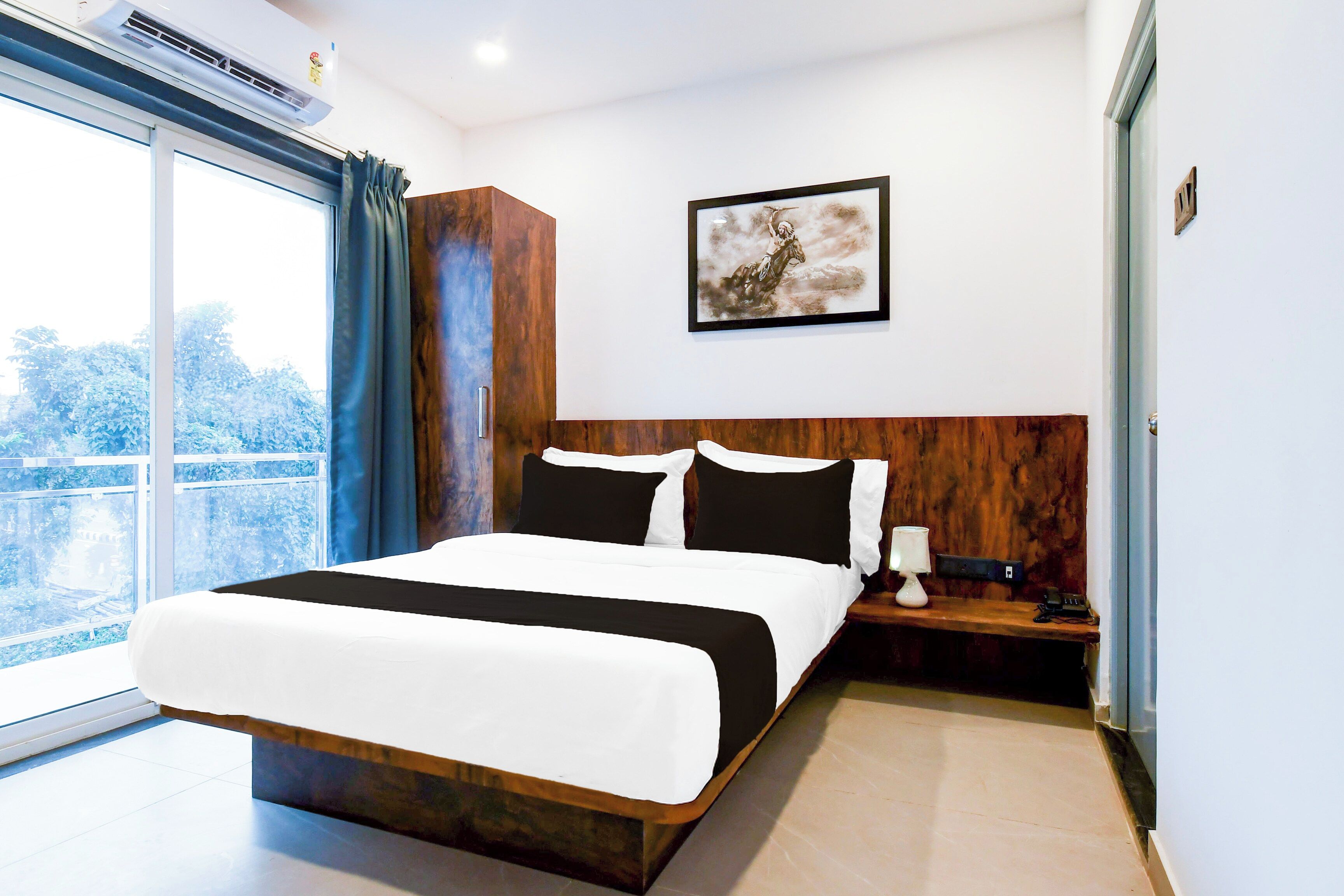 Une chambre d’hôtel avec un grand lit, une table de chevet, une lampe, un tableau encadré au mur et une vue sur les arbres par la fenêtre.