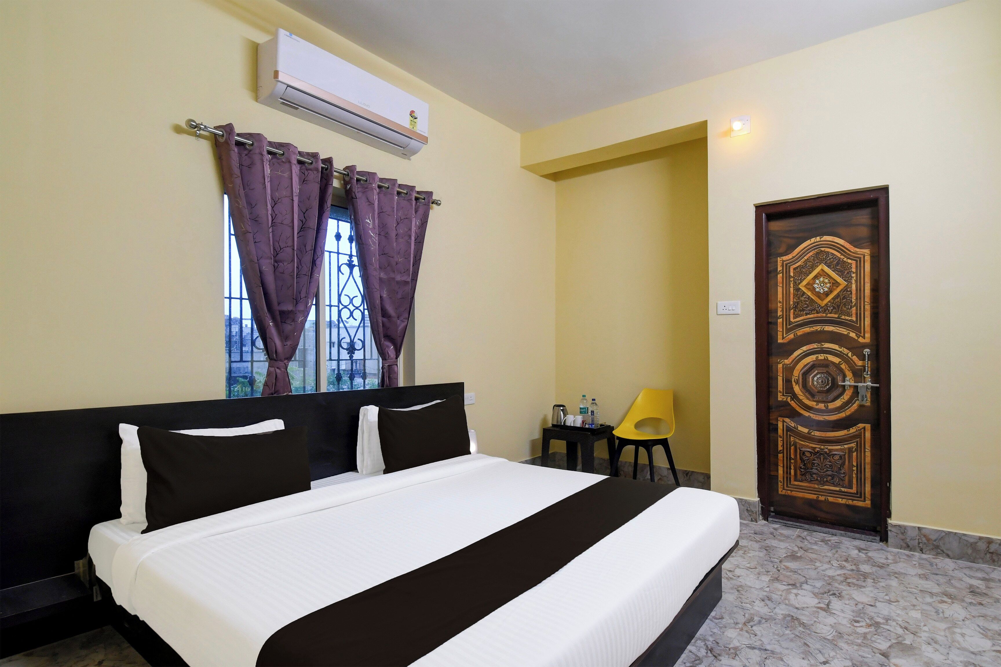 Habitación de hotel con cama, unidad de aire acondicionado, ventana con cortinas moradas, puerta con diseño decorativo y una pequeña mesa con una silla.