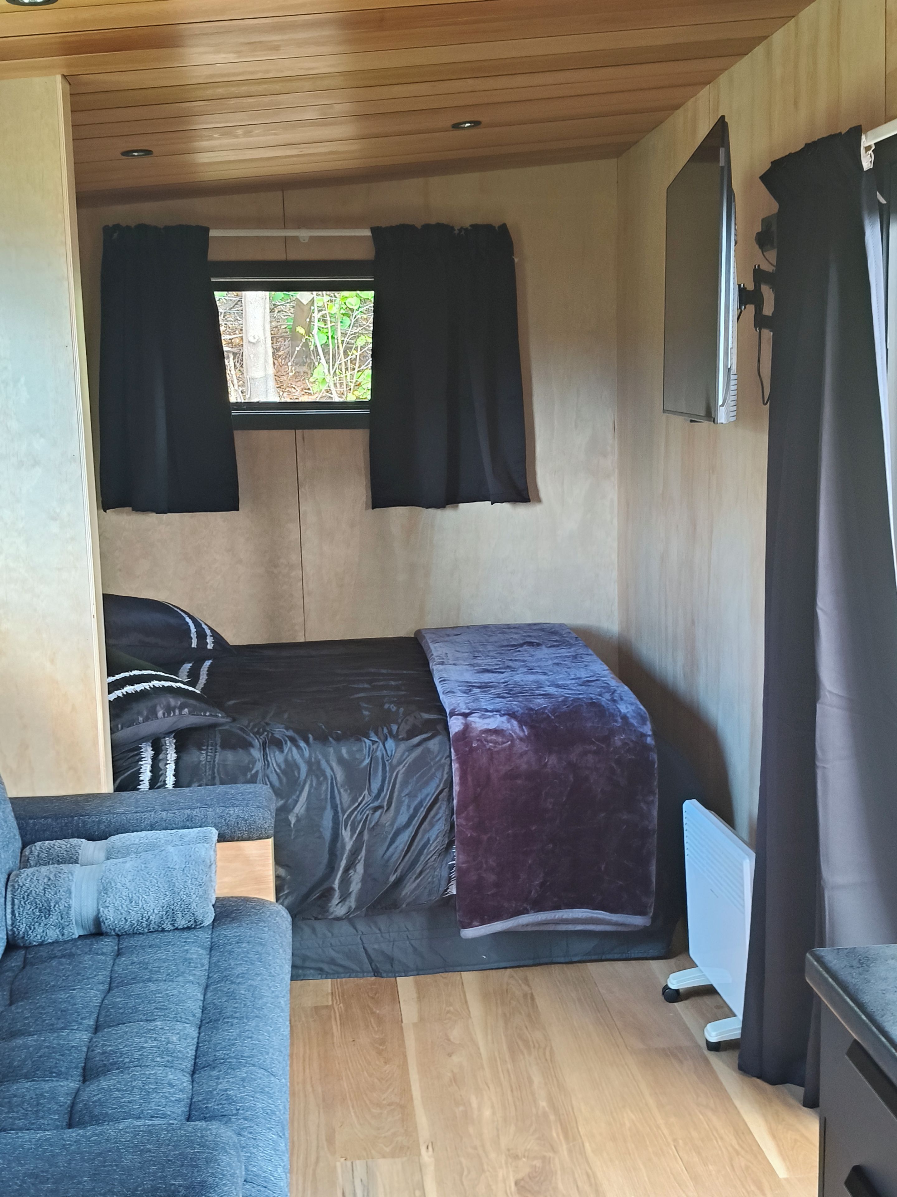 A compact room with a bed, a sofa, a window with curtains, and a mounted television.
