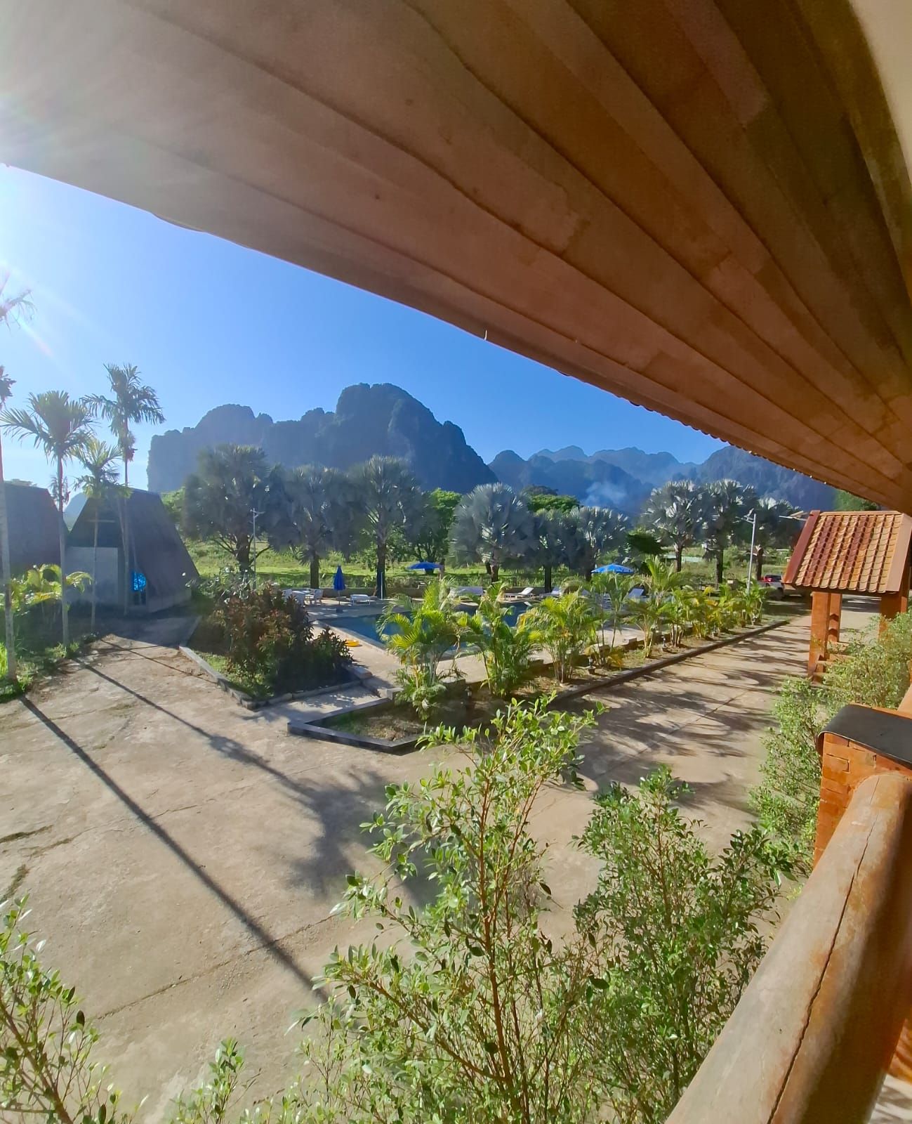 Une vue depuis un espace abrité donnant sur un complexe hôtelier, avec des palmiers, des chaises longues et une chaîne de montagnes en arrière-plan.