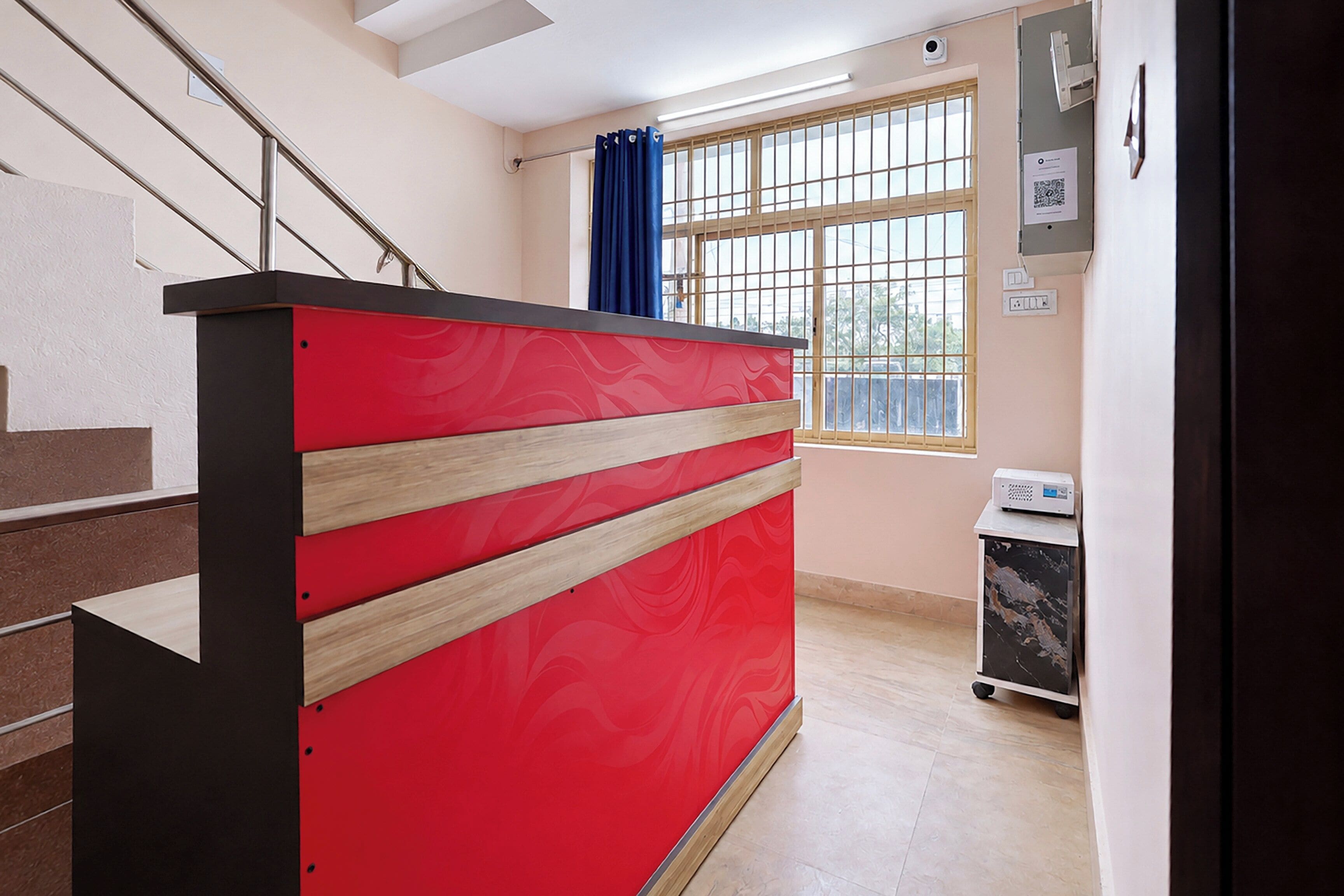 Una recepción con diseño rojo y negro, una escalera a la izquierda, un ventanal con cortinas azules y un carrito pequeño con un dispositivo encima.