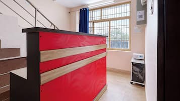 Una recepción con diseño rojo y negro, una escalera a la izquierda, un ventanal con cortinas azules y un carrito pequeño con un dispositivo encima.