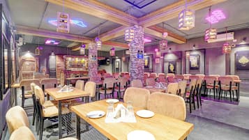 Interior de restaurante com várias mesas e cadeiras, letreiros de neon e um pilar de pedra.