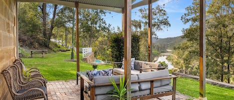 An outdoor patio with wicker furniture and a cushioned seating area, overlooking a river and greenery.
