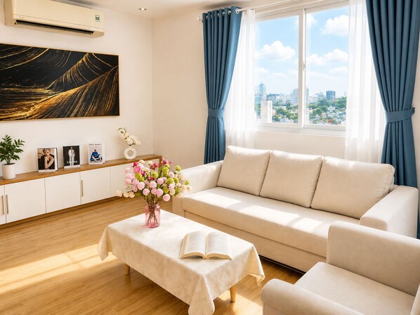 A modern living room with a sofa, a coffee table with an open book, a vase of flowers, and a city view through large windows. - Spacious 2BR Apt near Ben Thanh Market & D1 Free Wifi (Thành phố Hồ Chí Minh)