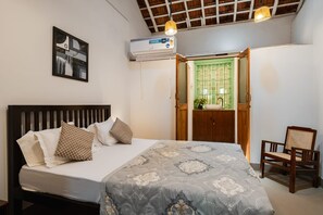 Un dormitorio con cama, almohadas, una mesita de noche, una silla, una ventana con vistas a un paisaje verde y un equipo de aire acondicionado.