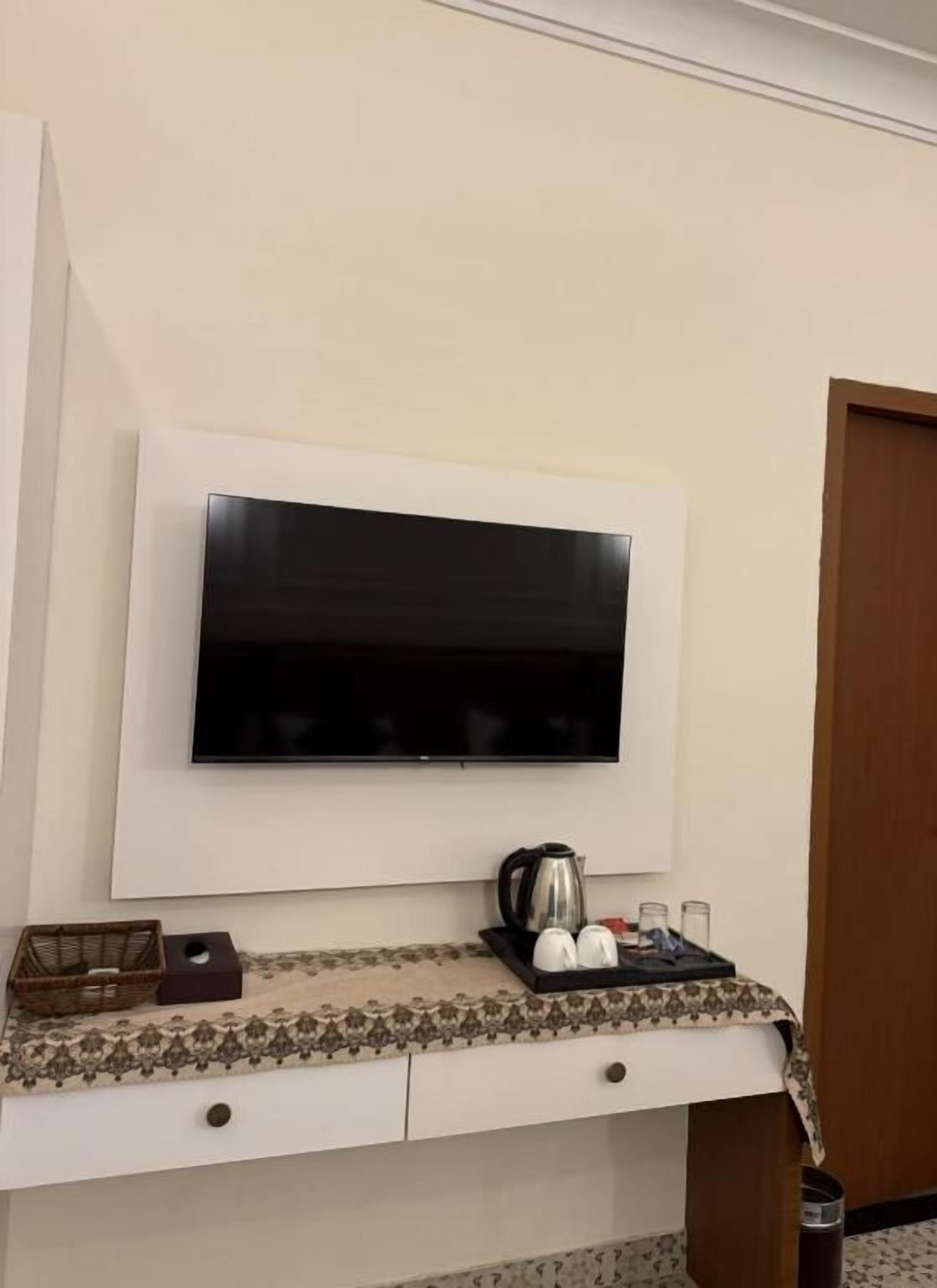 Televisão de tela plana presa na parede acima de uma escrivaninha com uma bandeja contendo chaleira, xícaras e uma caixa.