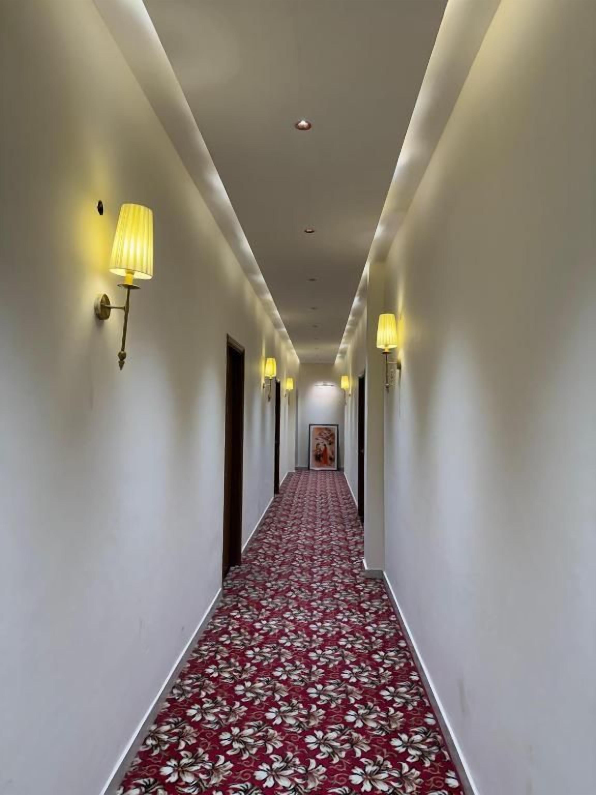 Corredor de hotel com tapete vermelho, luminárias na parede e um quadro emoldurado.