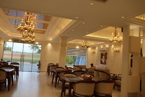 A spacious dining area with multiple tables and chairs, large chandeliers, and a view of the outdoors through large windows.