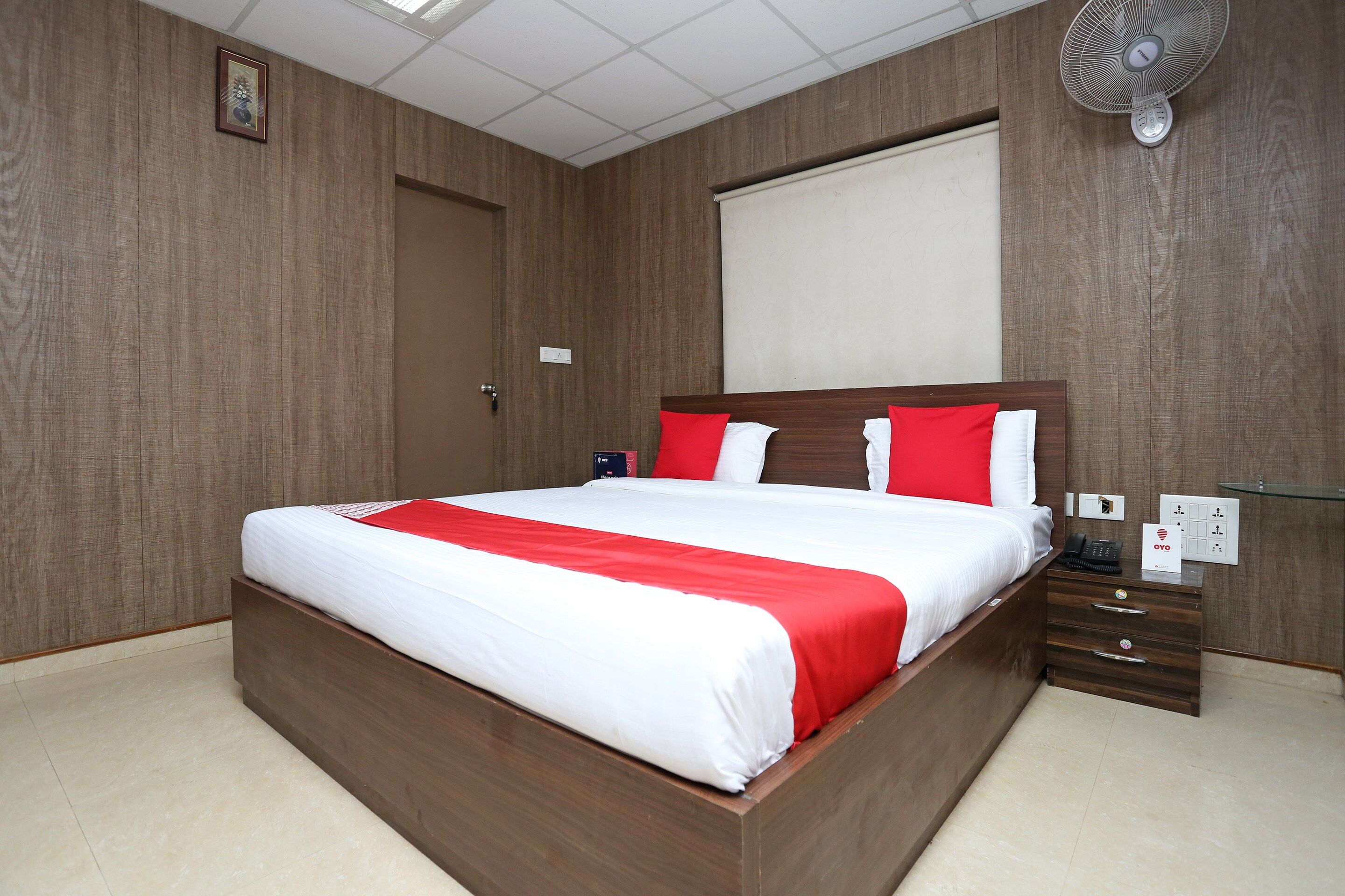 Ein Hotelzimmer mit einem hölzernen Bett, einem Nachttisch, einem Telefon und einem Wandventilator.