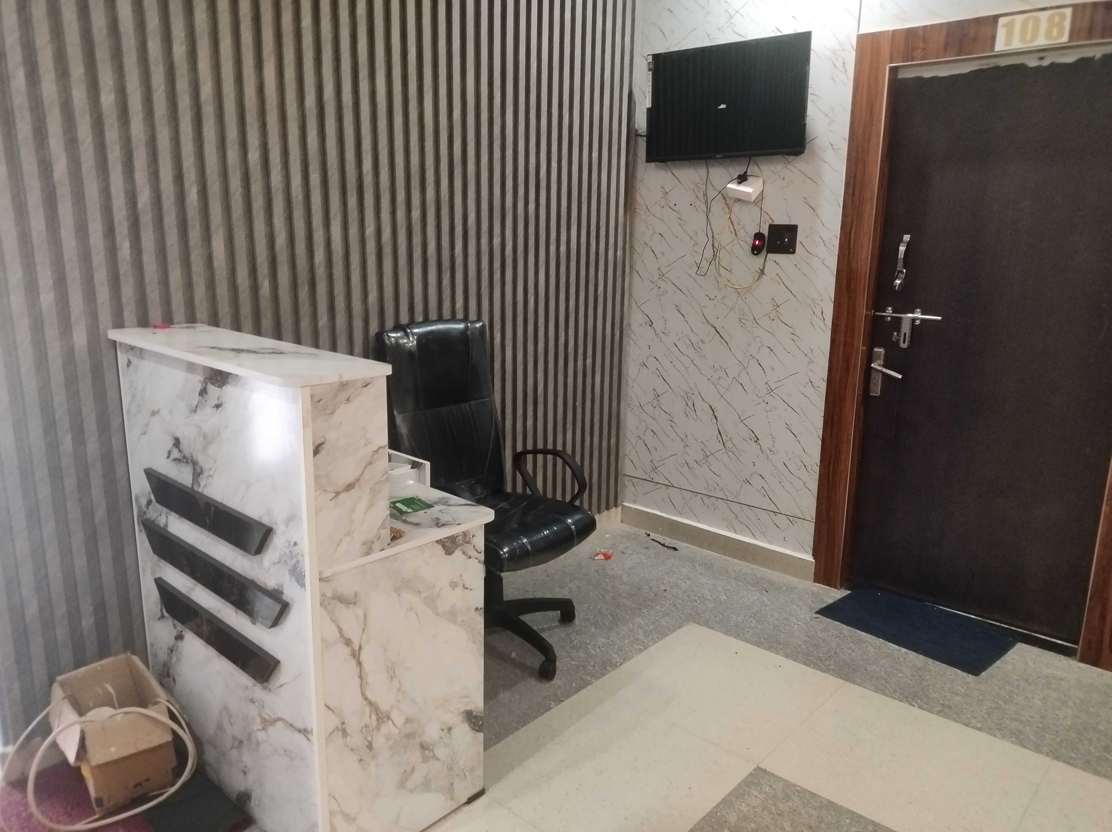Una habitación con un escritorio de mármol, una silla de oficina negra, un televisor montado en la pared y una puerta con una placa de número.