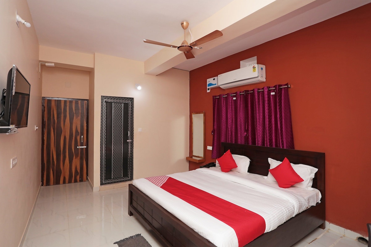 A hotel room with a bed, a ceiling fan, an air conditioning unit, a bathroom with a mirror, and a door.