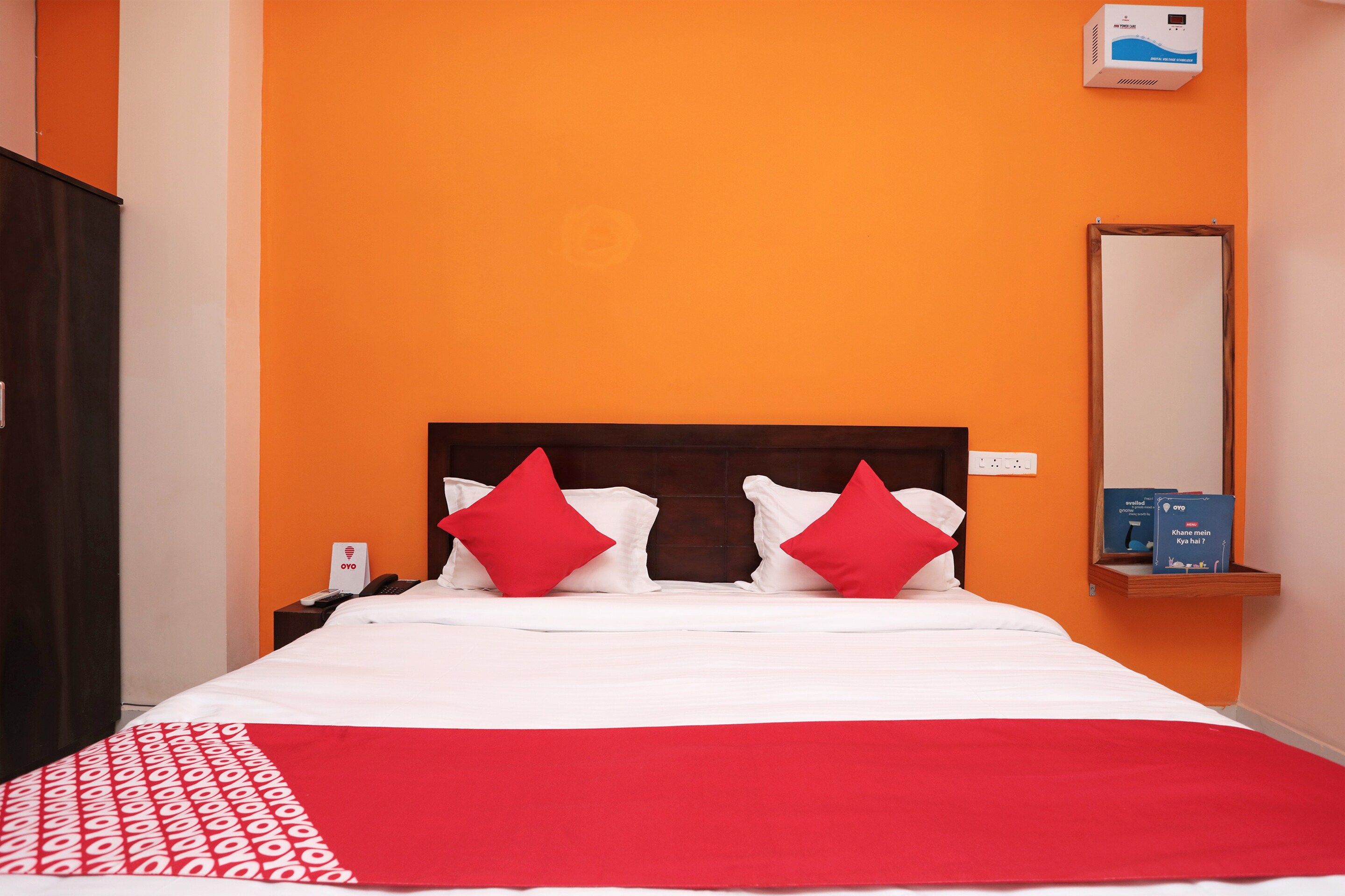 Habitación de hotel con pared naranja, cabecero de madera, cama con ropa blanca y almohadas rojas, mesita de noche con un teléfono y un reloj digital en la pared.