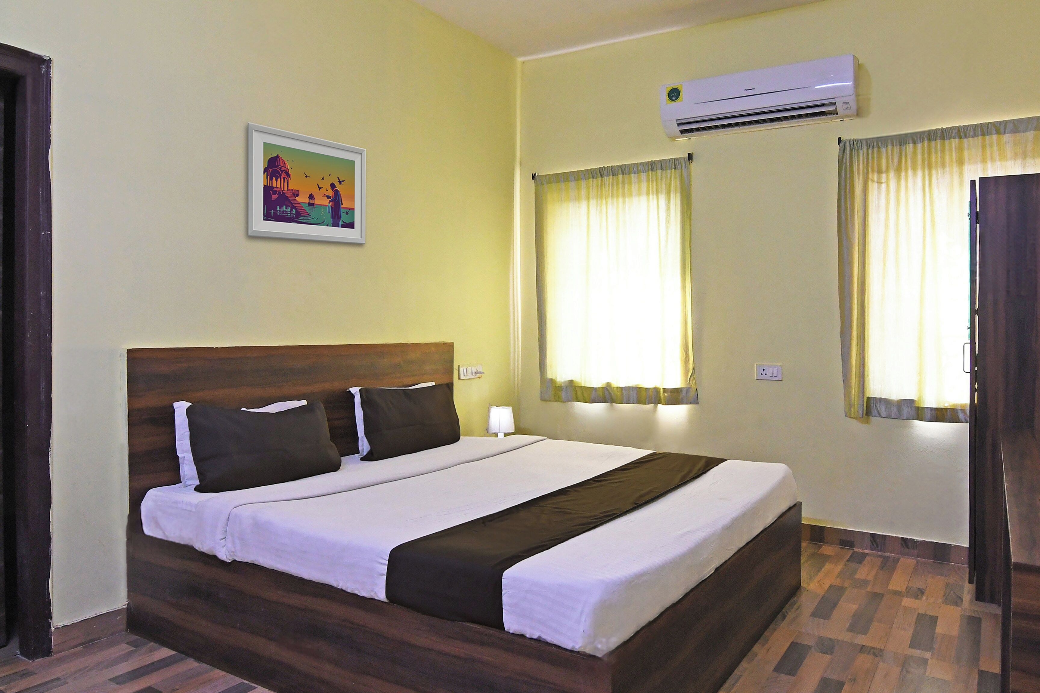 A hotel room with a bed, an air conditioner, a painting, and curtains.