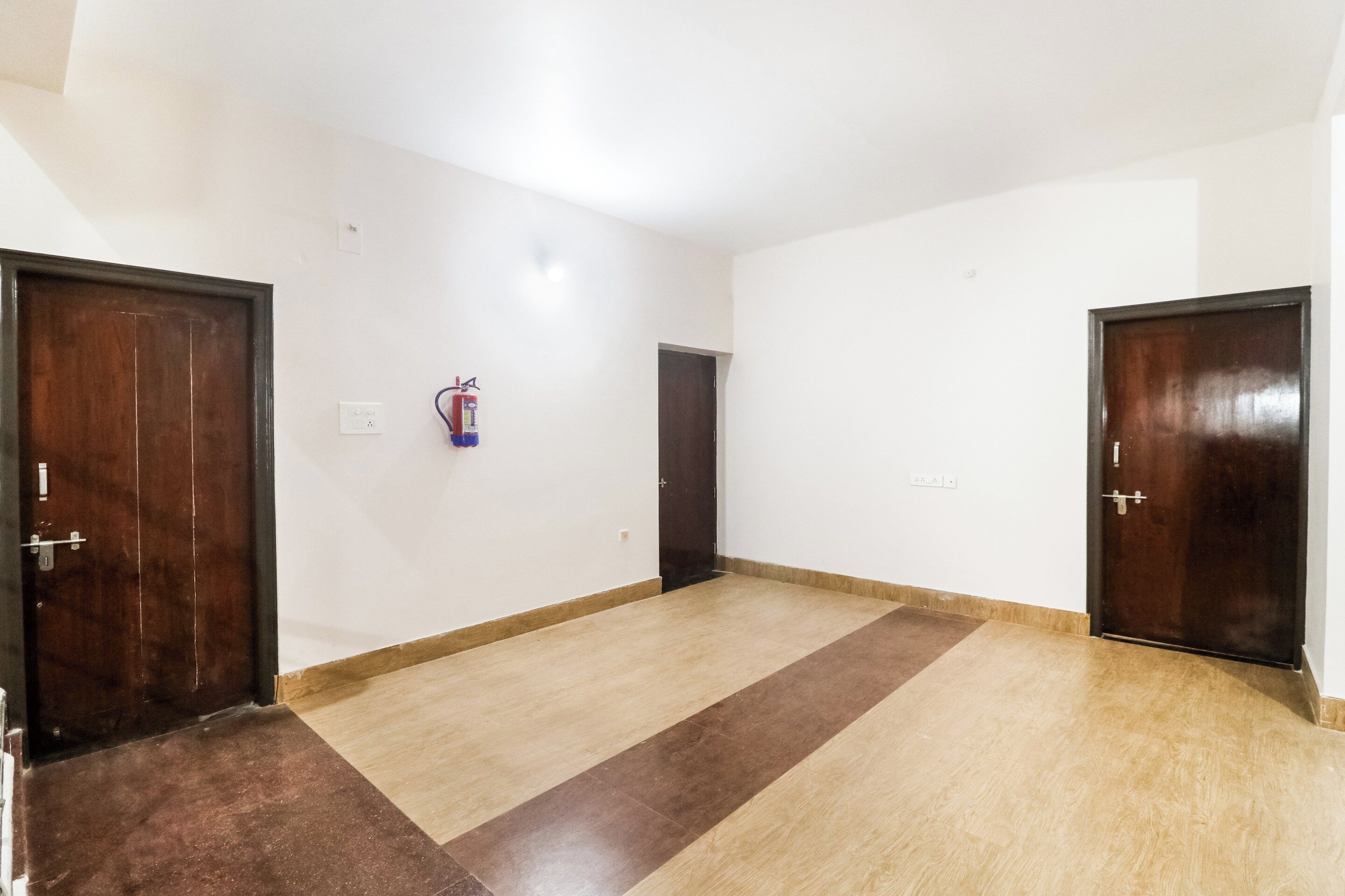 A hallway with two dark wooden doors, a fire extinguisher mounted on the wall, and a light switch.