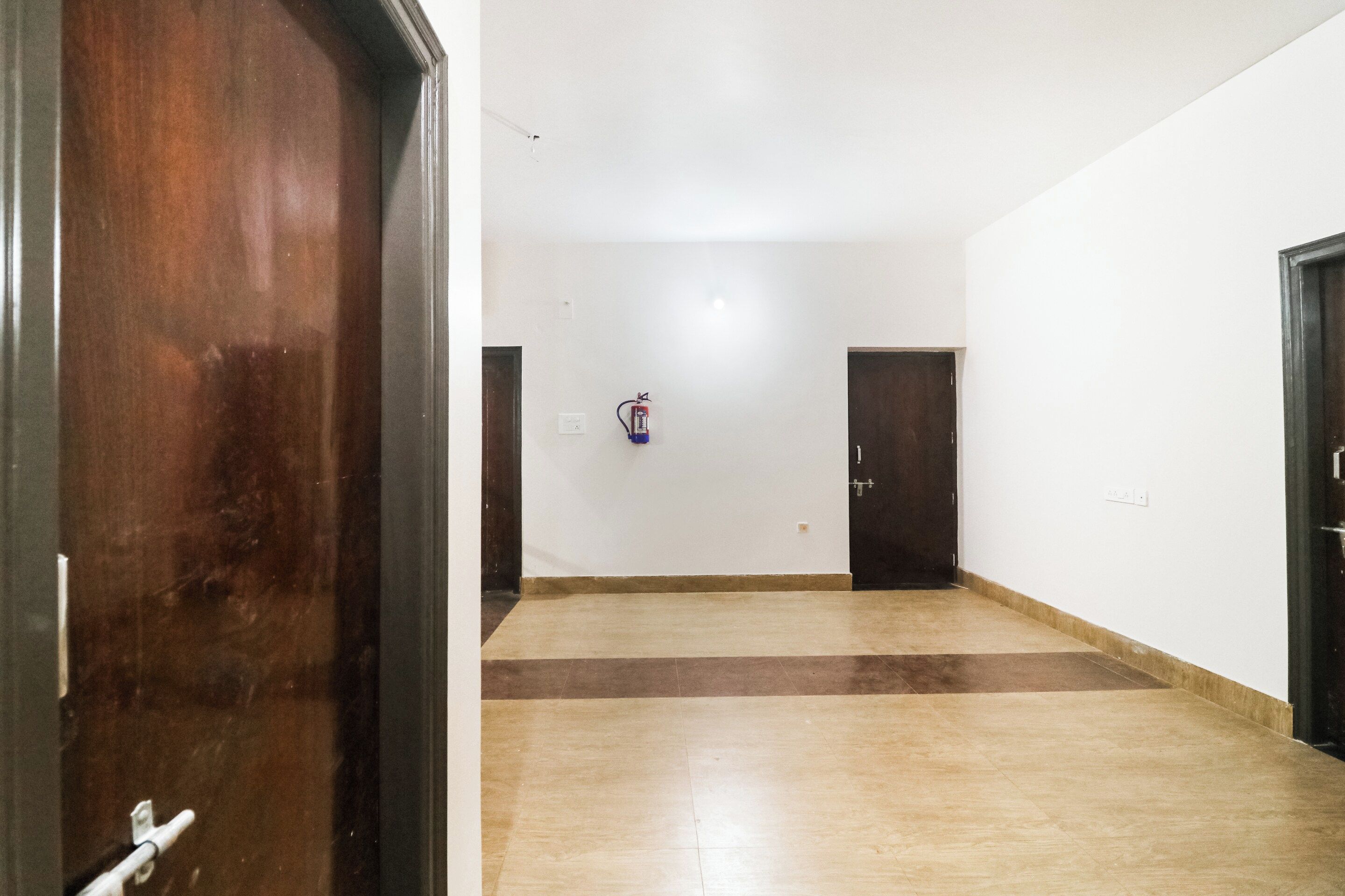 Un pasillo con una puerta de madera a la izquierda, un detector de incendios en la pared y dos puertas cerradas a la derecha.