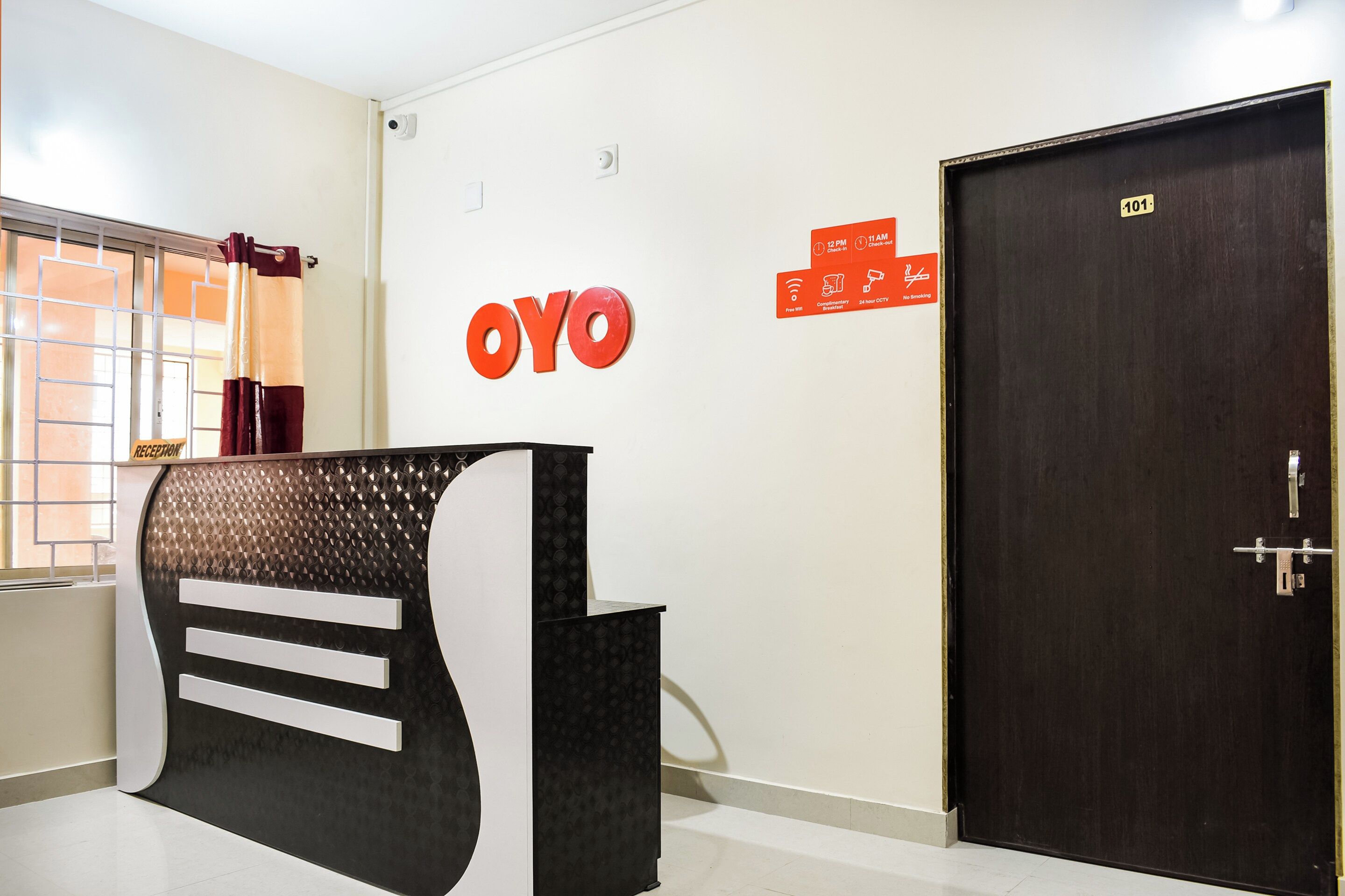 Un área de recepción con mostrador, un letrero en la pared con la marca 'OYO', una puerta con el número '101' y una ventana con cortina.