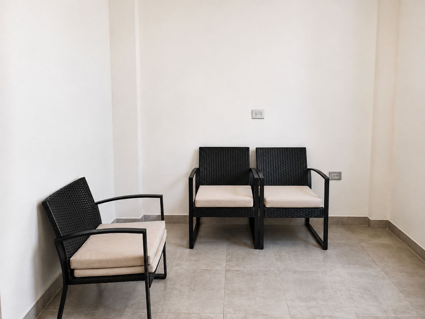 A corner of a room with two black wicker chairs with beige cushions, a white wall, and a tiled floor.