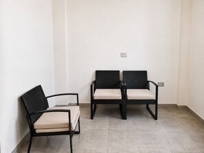 En hörna i ett rum med två svarta fåtöljer i rotting med beige kuddar, en vit vägg och ett klinkergolv.
