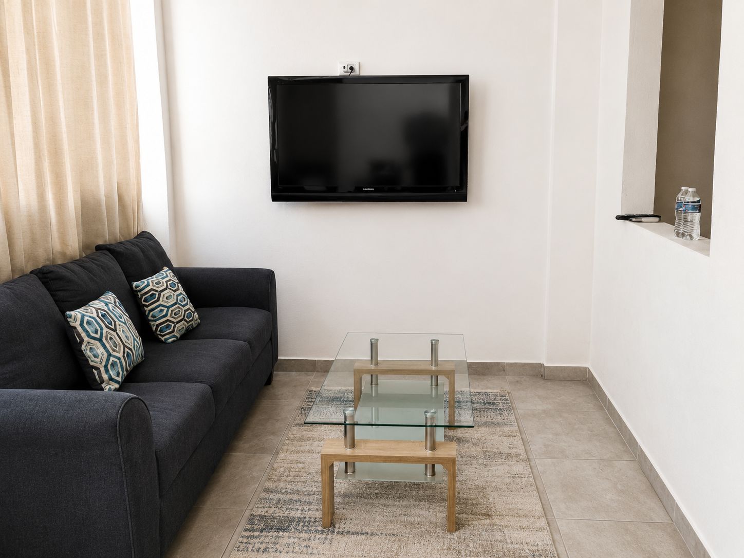 A modern living room with a black sofa, patterned cushions, a glass coffee table, and a flat-screen TV mounted on the wall.