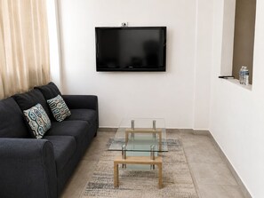 Ett modernt vardagsrum med en svart soffa, mönstrade kuddar, ett soffbord i glas och en platt-TV som är monterad på väggen.