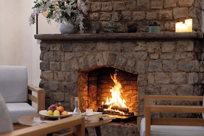 Una chimenea de piedra con fuego, un florero con flores, y una mesa con fruta y una vela.