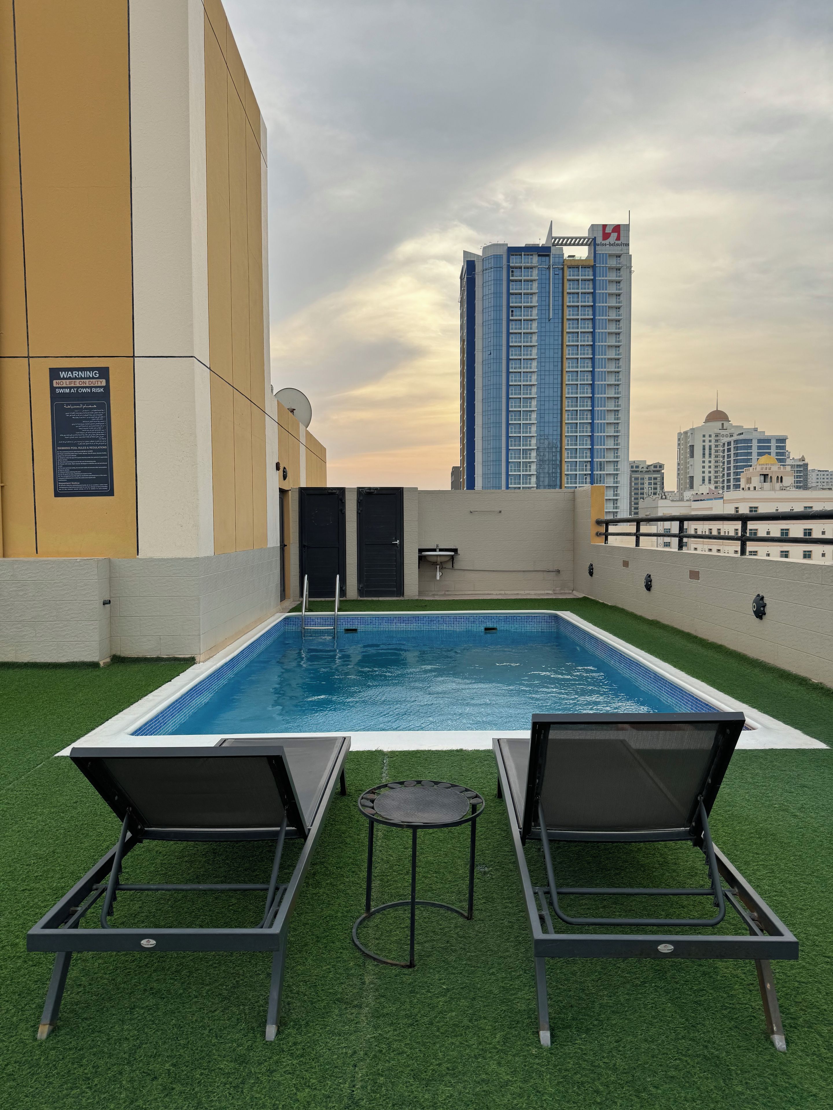 A rooftop pool with two lounge chairs and a small table, surrounded by artificial grass, with a city skyline in the background.