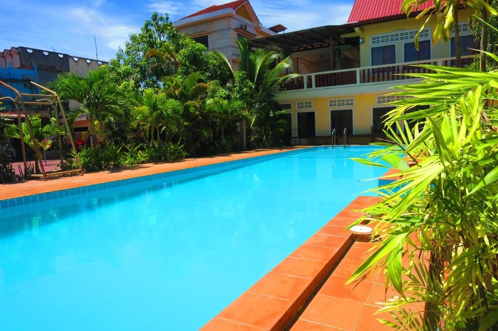 A swimming pool with a clear blue water surface, surrounded by lush green tropical plants and a building with a red roof in the background.