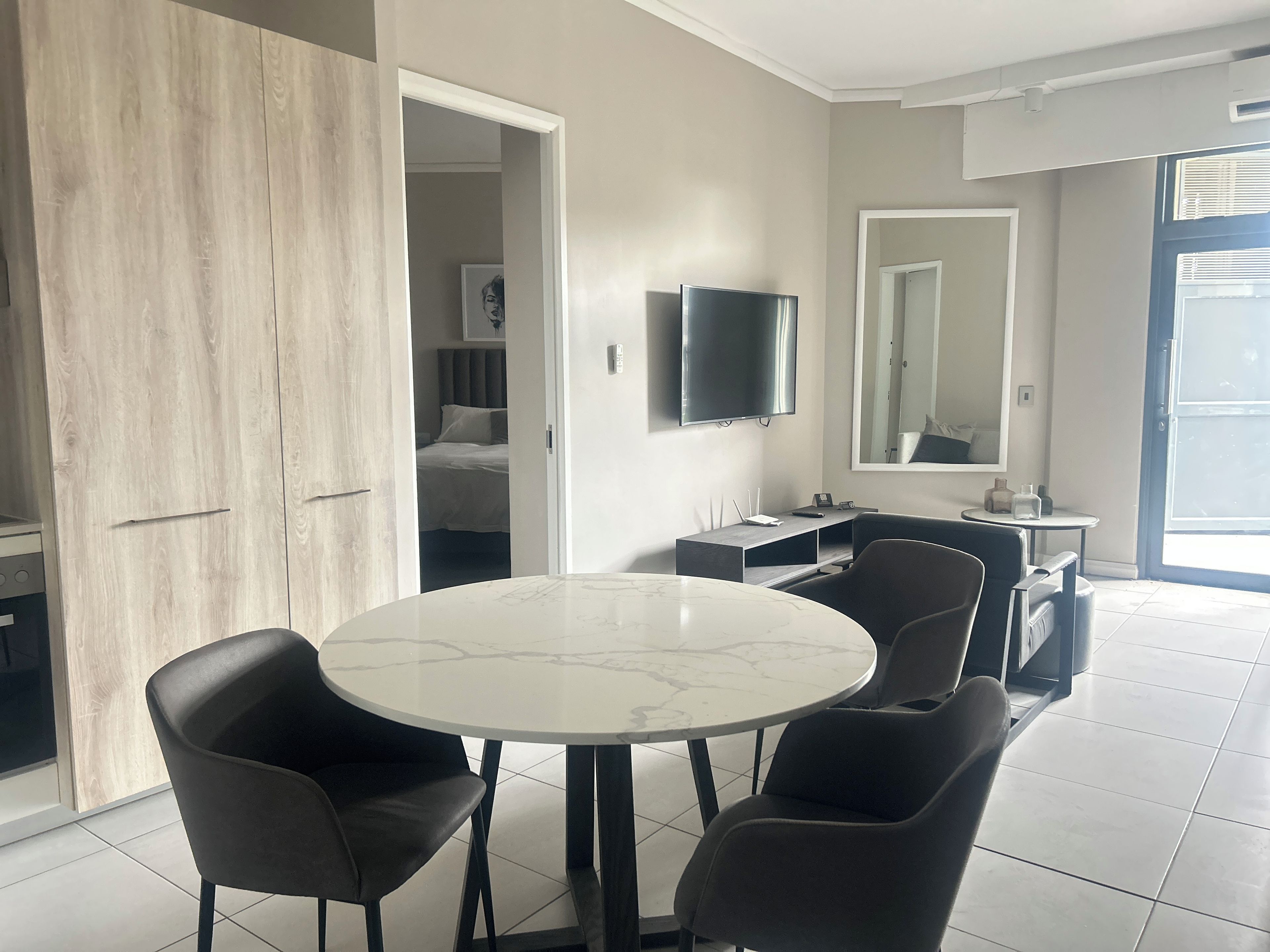 A modern hotel room with a round dining table, four chairs, a flat-screen TV, a mirror, and a wardrobe.