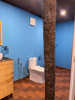A bathroom with a blue wall, a wooden vanity, a toilet, and a tree trunk in the center. - The Racha Agonda (Agonda)