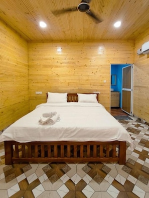Una habitación con paneles de madera, una cama, un ventilador de techo y un baño visible a través de una puerta abierta.
