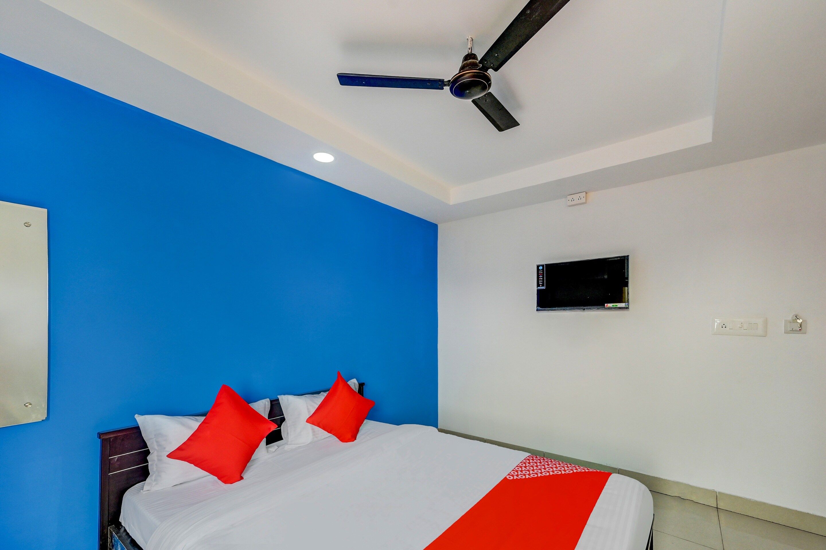 A room with a bed, a ceiling fan, a wall-mounted TV, and a power outlet.
