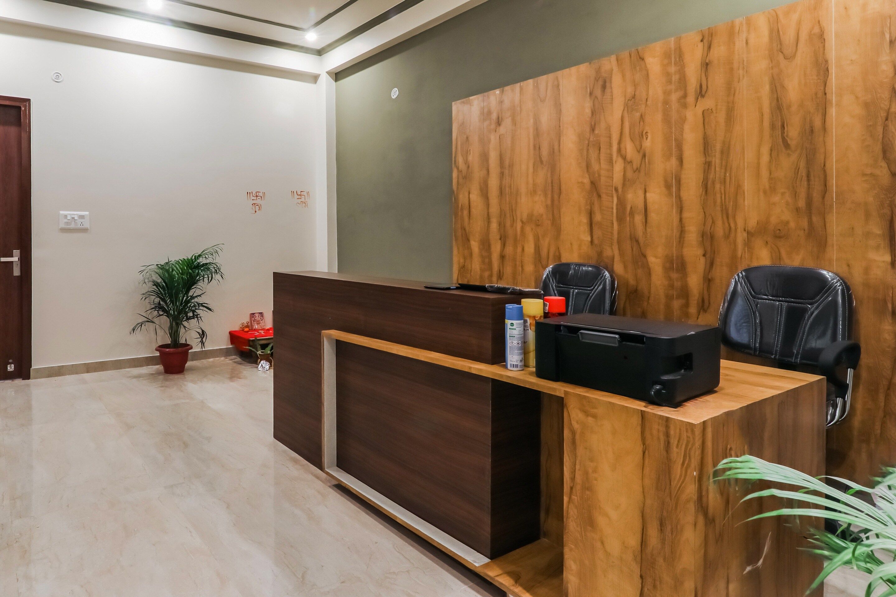 A reception desk with a wooden finish, two black chairs, a potted plant, and a red table with a small orange object.