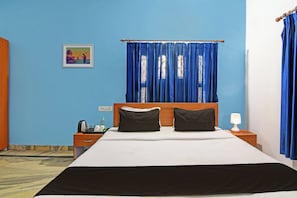 A hotel room with a bed, two pillows, a nightstand with a lamp, a picture on the wall, and blue curtains. - Hotel O Home Elite Stay Near Shri Shri (Bhubaneswar)