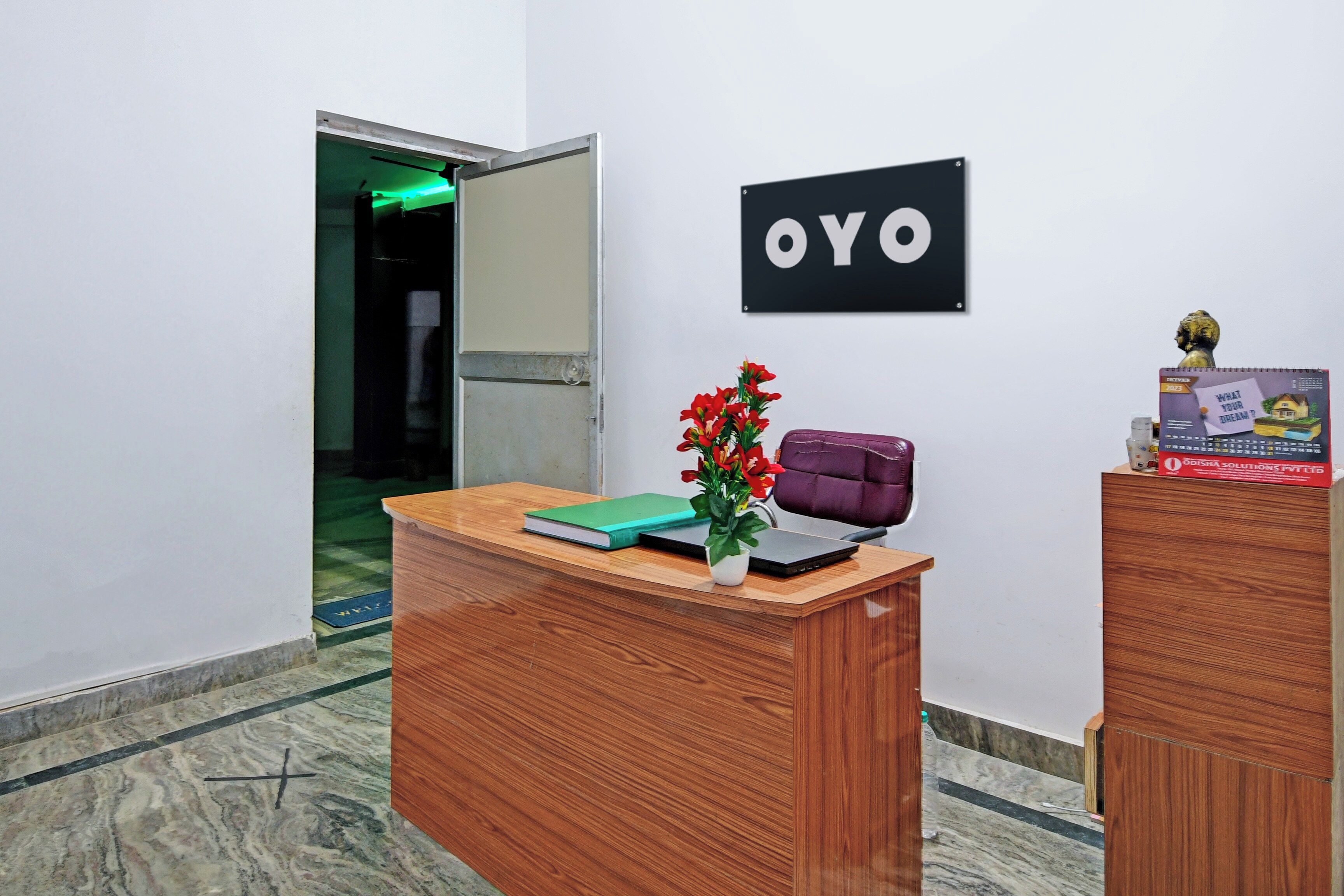 A reception desk with a sign that reads 'OYO' on the wall, a potted plant, a purple bag, and a calendar on the desk.