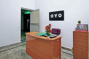 A reception desk with a sign that reads 'OYO' on the wall, a potted plant, a purple bag, and a calendar on the desk. - Hotel O Home Elite Stay Near Shri Shri (Bhubaneswar)