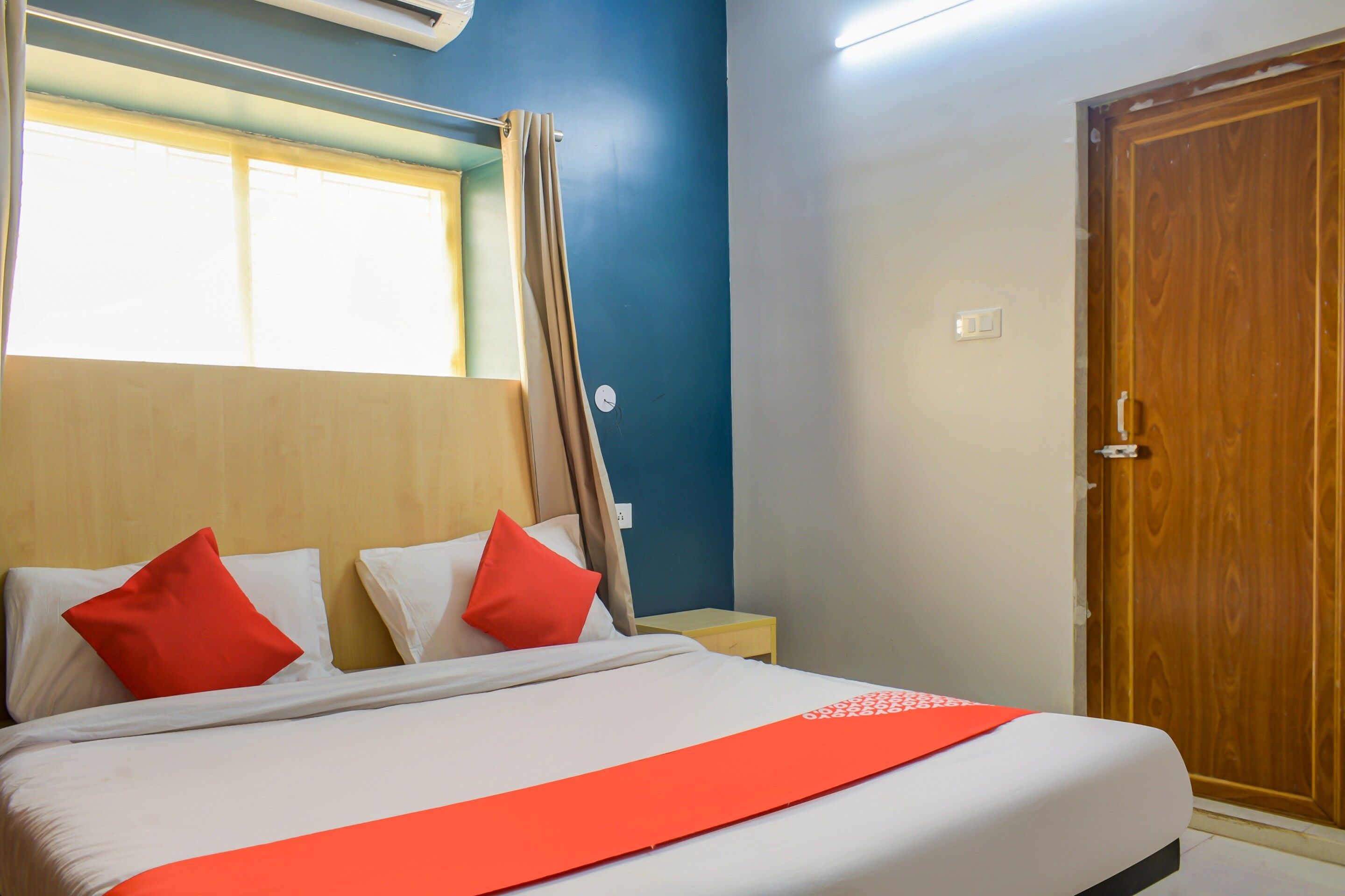 A hotel room with a bed, two pillows, a nightstand, a wooden door, and a window with curtains.