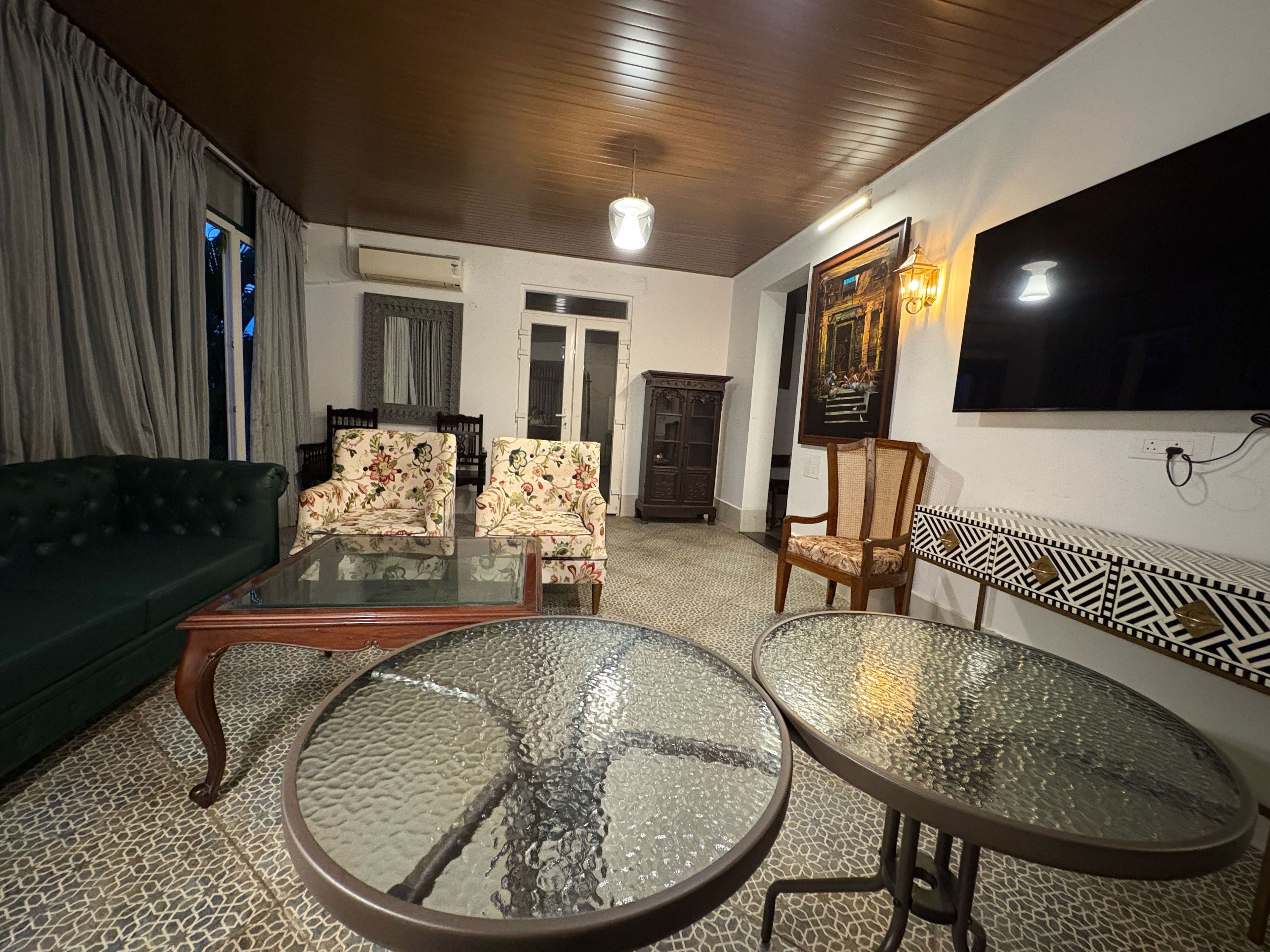 Una sala de estar con un sofá verde, dos sillones estampados, una mesa de centro de vidrio, un armario de madera y un televisor en la pared.