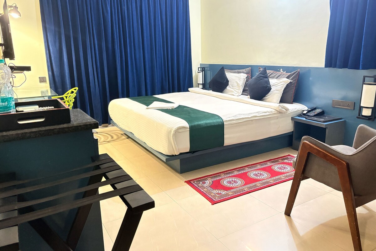 A hotel room with a bed, bedside tables, a desk with a chair, a red patterned rug, and a window with curtains.
