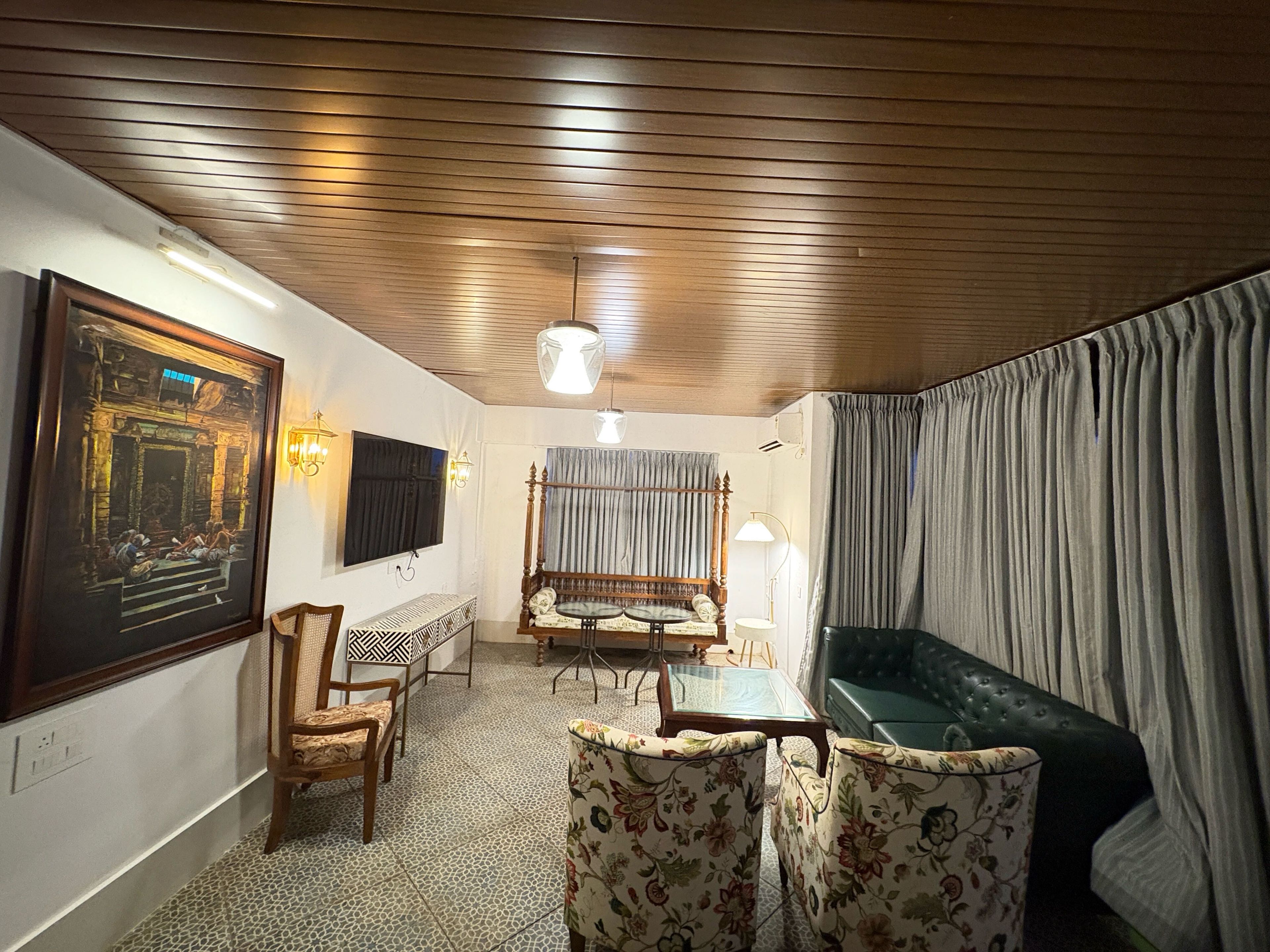 A room with a wooden ceiling, a television mounted on the wall, a sofa, a patterned floor, and a framed painting.