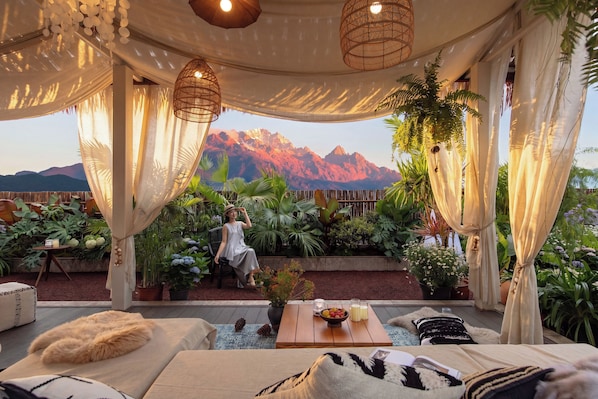 A cozy outdoor seating area with a view of mountains, surrounded by lush plants and draped fabric.