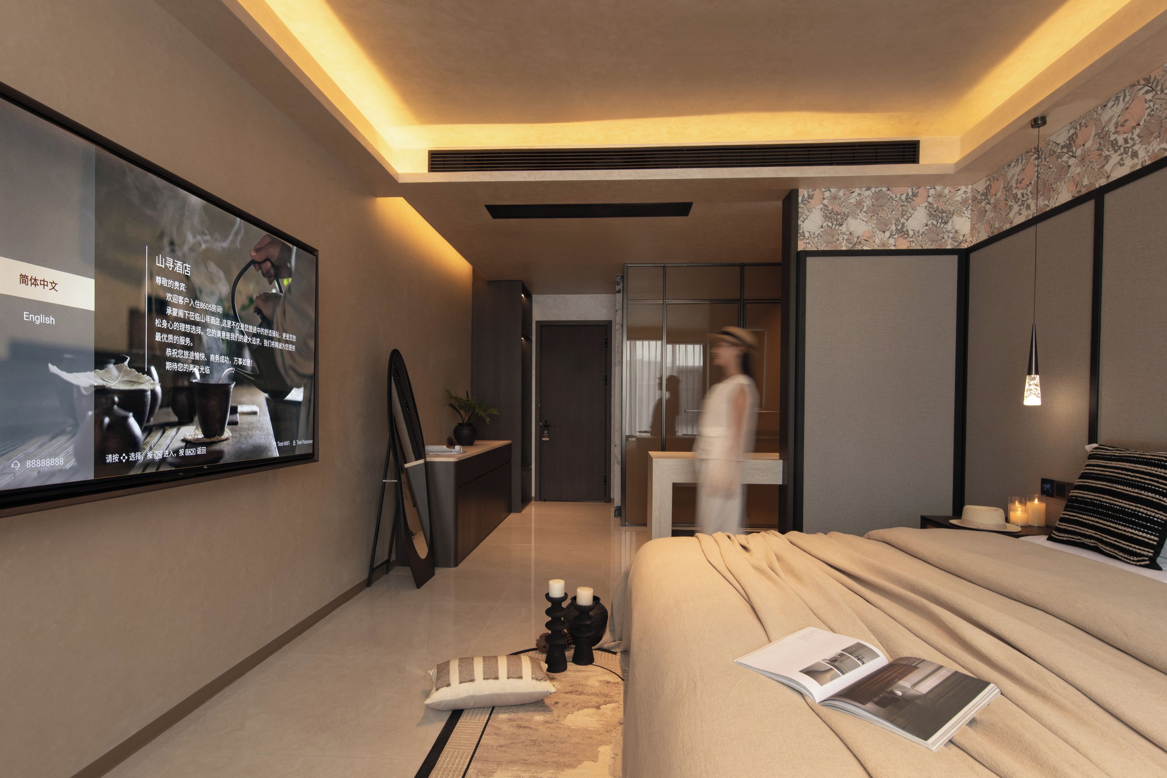 A modern hotel room with a large flat-screen TV, a bed with a magazine, a small table with candles, and a mirror.