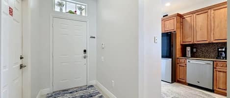 A hallway with a door, a small window with plants, and a kitchen area with wooden cabinets and a dishwasher.