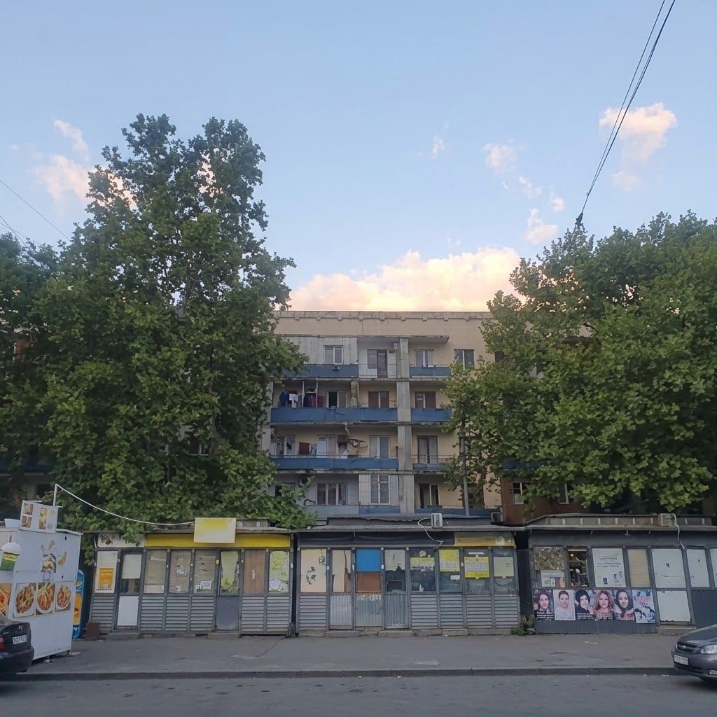 A multi-story building with balconies, a storefront with closed shutters, and trees in the foreground.