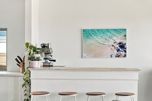 A kitchen with a coffee machine, bar stools, and a wall-mounted painting of a beach scene. - The Ultimate Oceanfront Entertainer (Geographe)