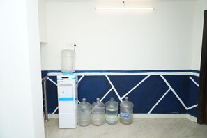 Ein Wasserspender mit einem großen Wasserkrug und mehreren kleinen Flaschen auf einem Kachelboden, vor einer Wand mit blau-weißem geometrischen Muster.